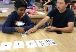 Two students using playing cards at a table with cards showing differing number of dots laying on the table in front of them.