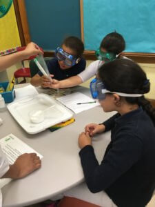Students wearing goggles sitting at a table looking observe a chemical reaction.