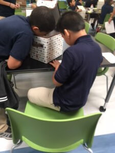 One student taking notes, while the other is looking inside a box.