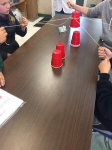 A table is shown with students using string to stack red solo cups.
