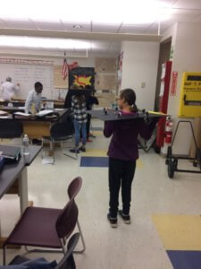 A student stands with the moon phase board over her head while other students look work in the background.