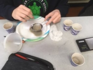 A student's hands are shown sifting a mixture onto a paper plate surrounded by small paper cups, a funnel and a scale.