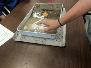 A paint tray is shown that contains a model beach with sand and water while a hand holds an empty water bottle over the water.