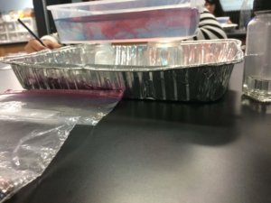 A container of water that has blue and red food coloring representing storm fronts sitting on jars in a foil tray.